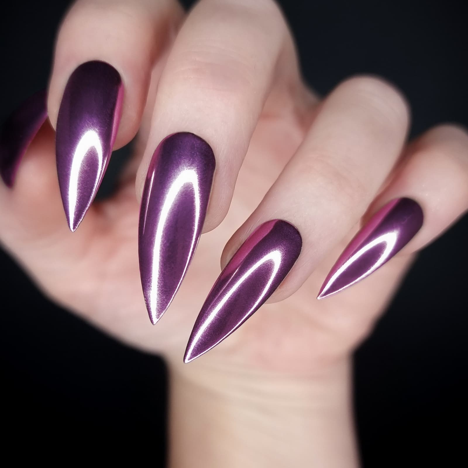 Close-up of a hand displaying stiletto-shaped chrome metallic purple gel nails against a black background