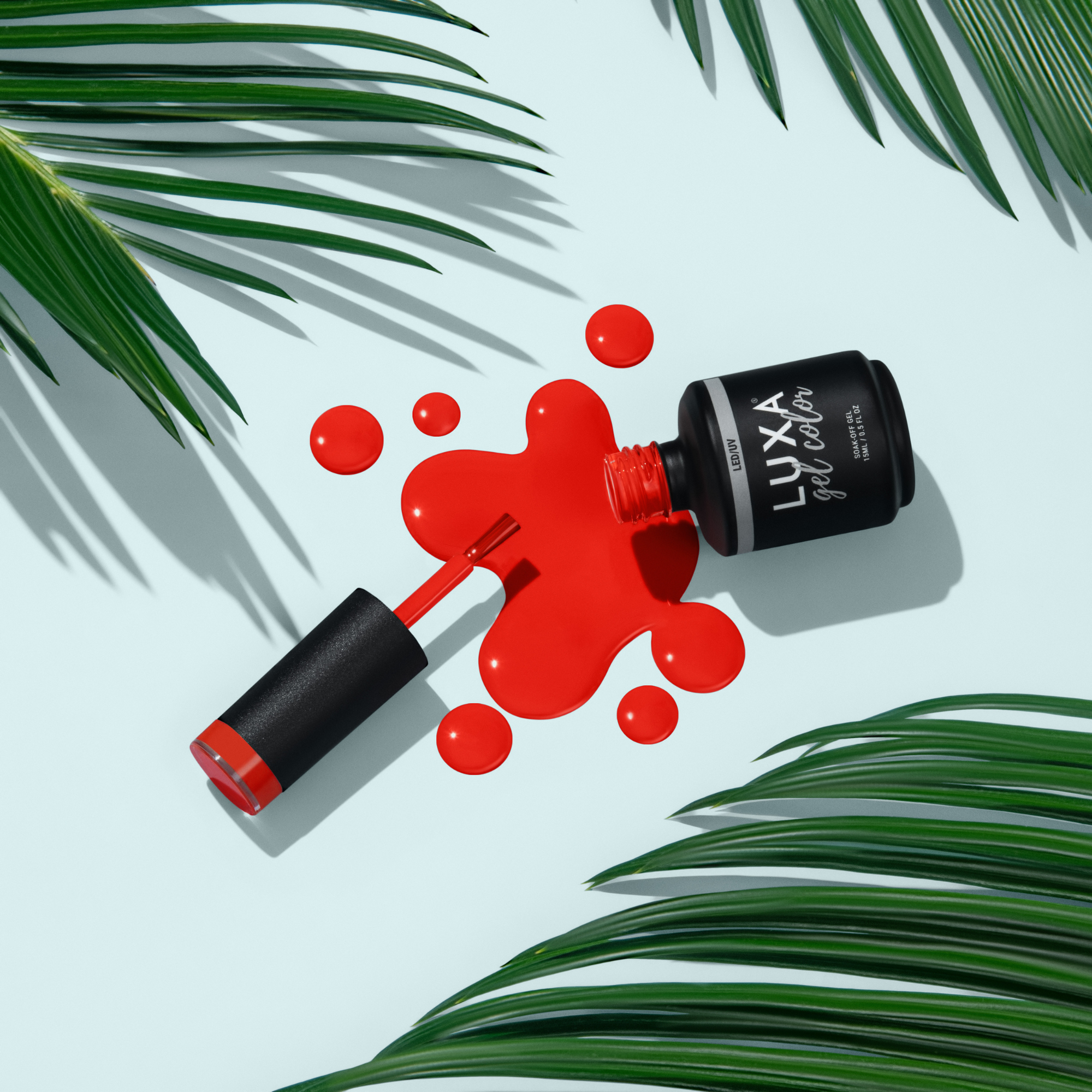 Top view of a red gel polish bottle and brush with red polish drops on a pale blue surface surrounded by palm fronds