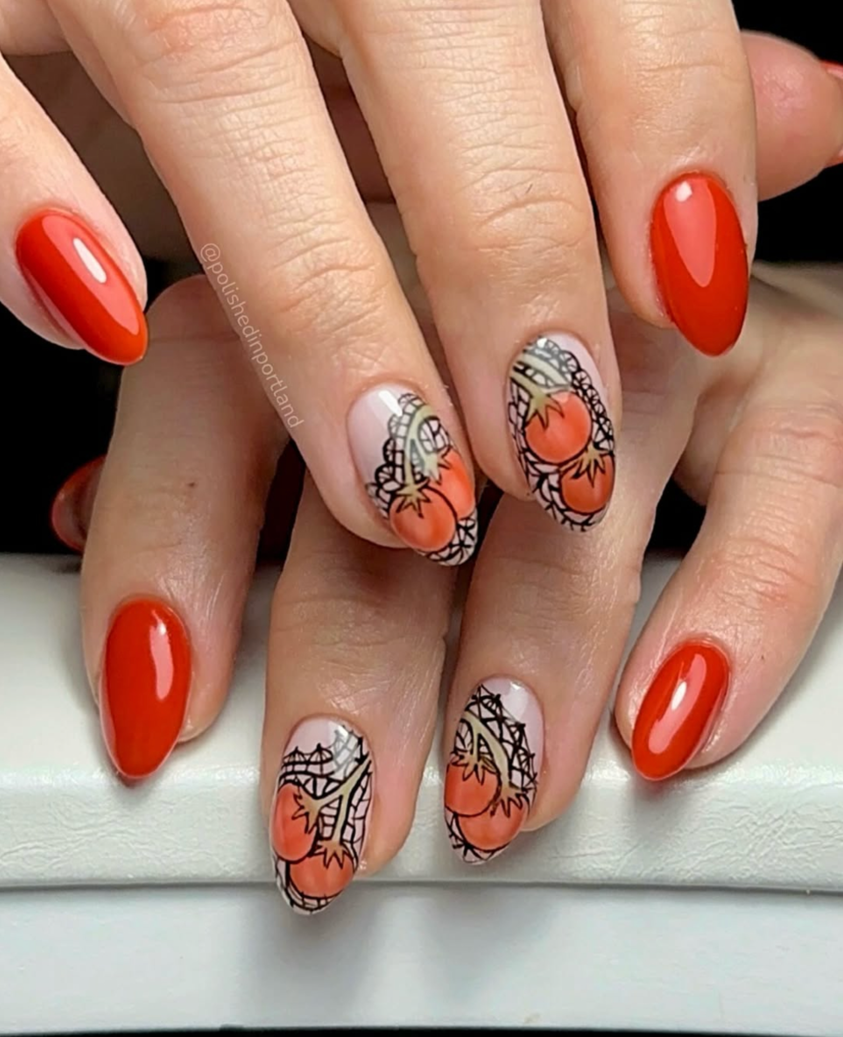 Close-up of hands showing glossy tomato-red almond nails and accent nails with tomato fruit and black lace nail art on a pale pink base