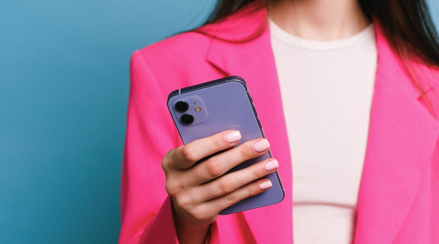 Woman wearing a pink blazer holding a lavender smartphone, showing pale pink manicured nails against a teal background