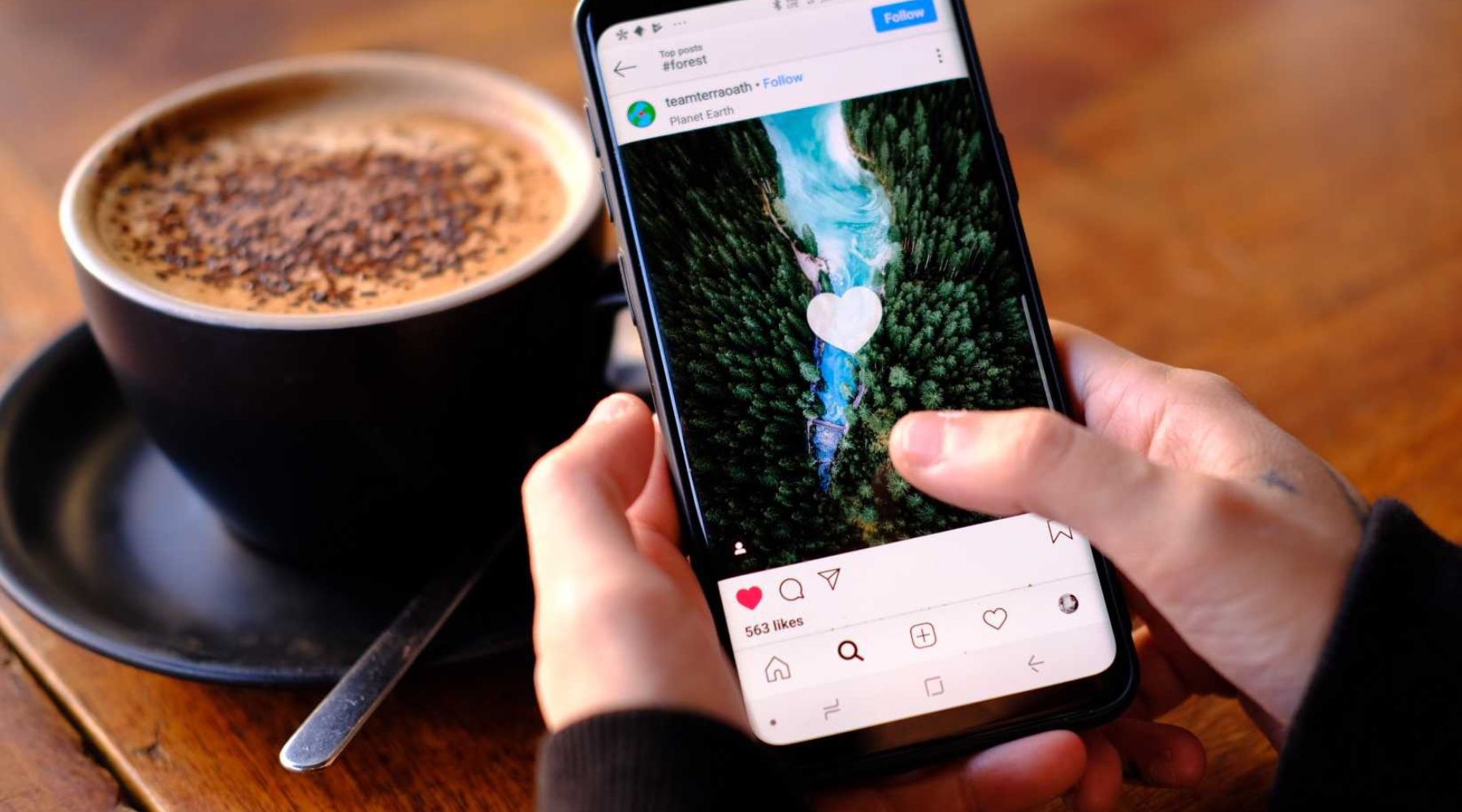Person holding smartphone tapping like on an Instagram photo of a river through a forest, cappuccino on a wooden table