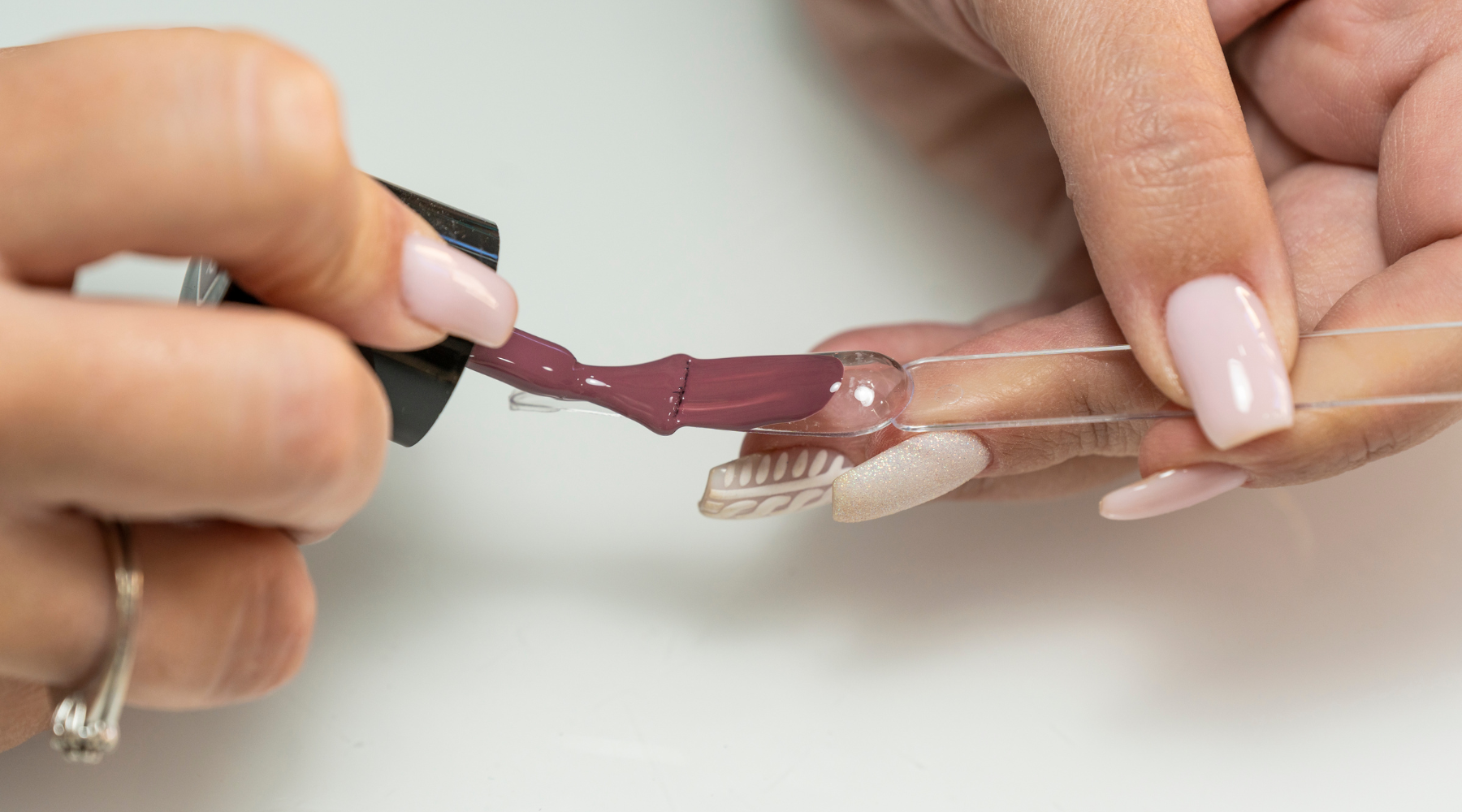 Close-up of hands applying mauve gel nail polish to a clear nail tip swatch on a white background