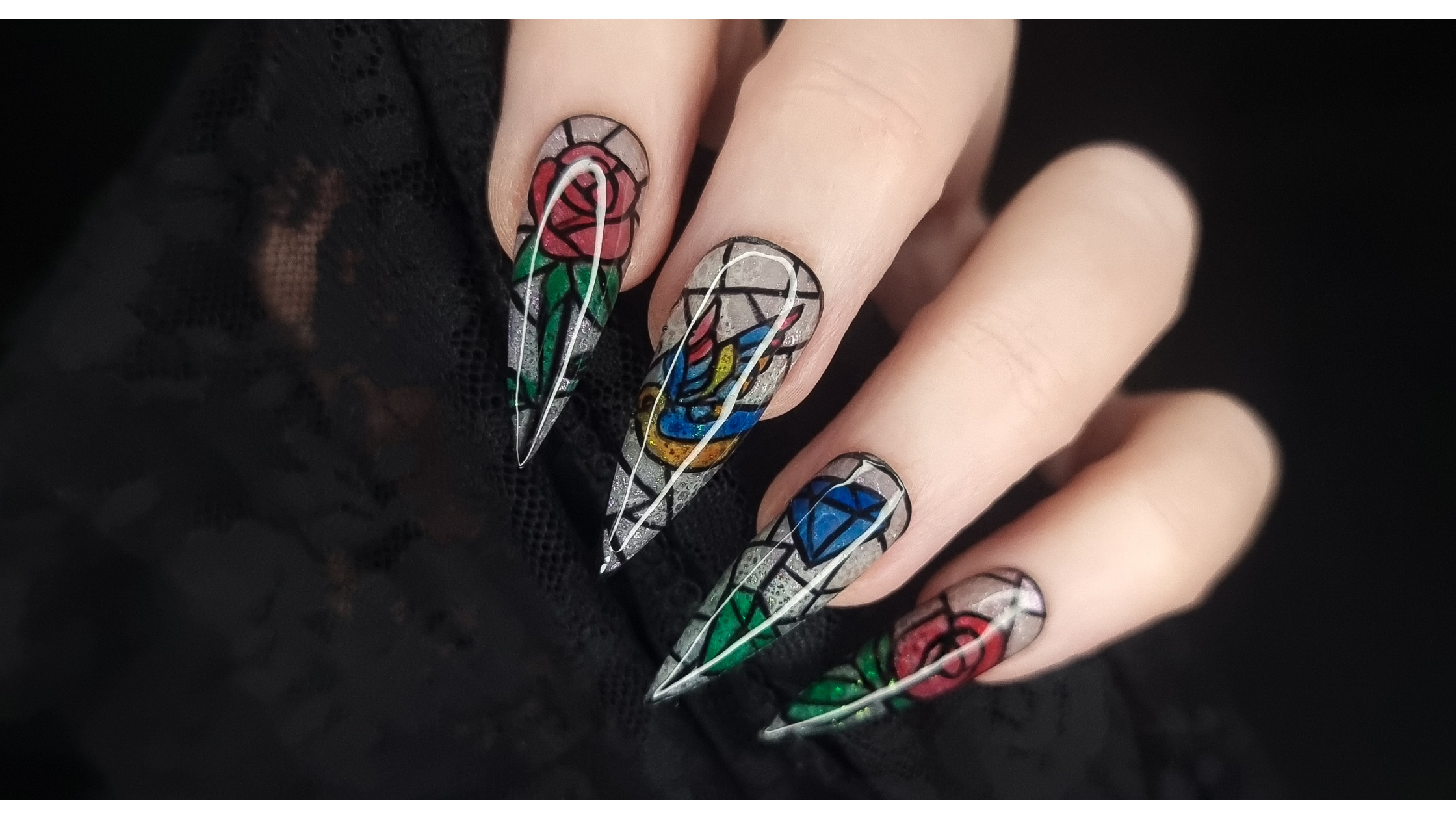 Hand with long stiletto nails showing stained-glass rose, diamond, and wing designs on silver glitter tips against a black lace background