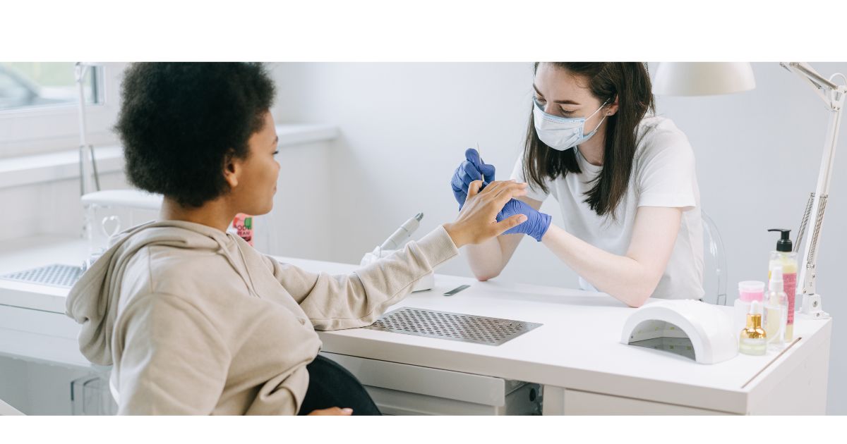 Nail technician wearing a mask and gloves filing a client’s nails at a white salon workstation with manicure tools and an LED lamp