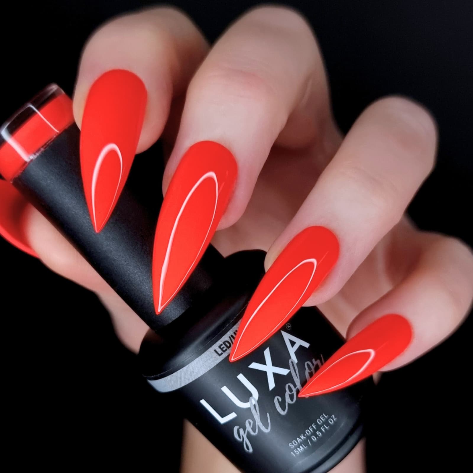 Hand holding a LUXAPOLISH gel polish bottle showing long matte red stiletto nails with white oval accents on a black background