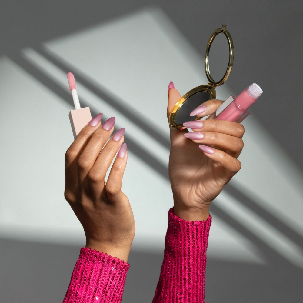 Hands with long pink ombre gel nails holding a lip gloss wand and an open compact mirror against a gray background with diagonal shadows