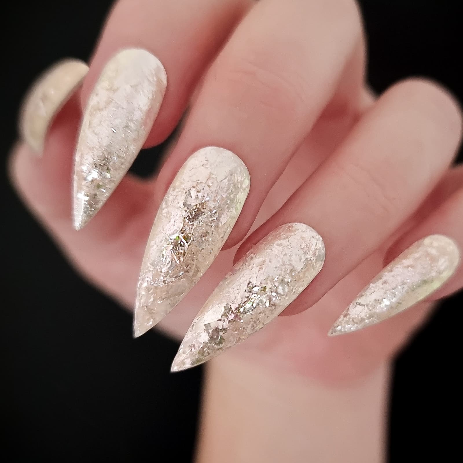 Close-up of stiletto gel nails showing textured pale pink-white polish with iridescent flakes against a dark background