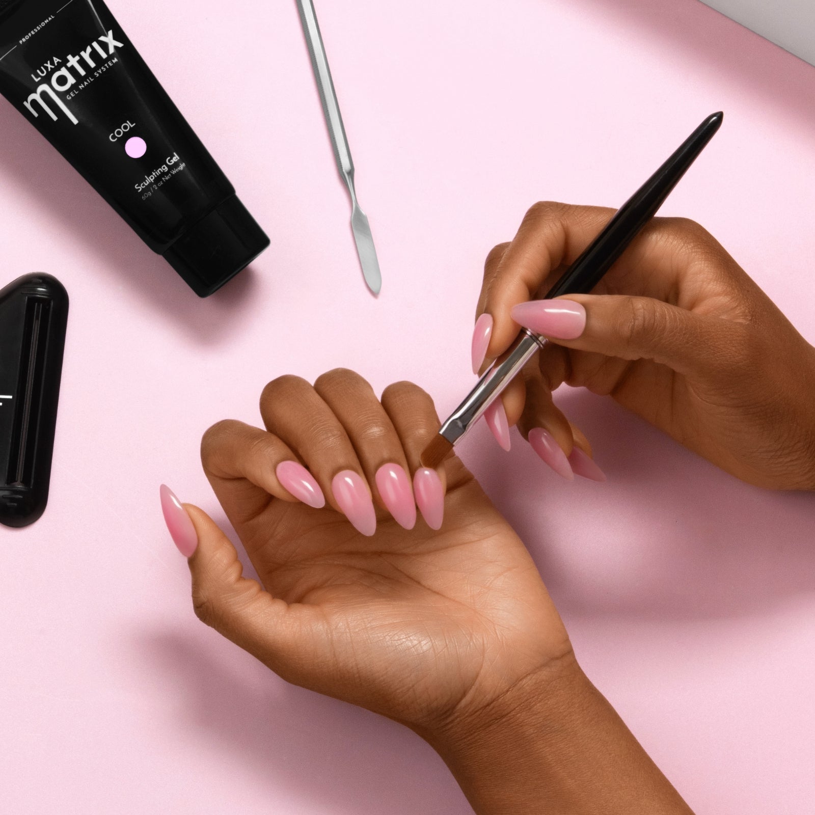 Hands applying pink almond gel nails with a brush on a pale pink background, Matrix gel tube and manicure tools nearby
