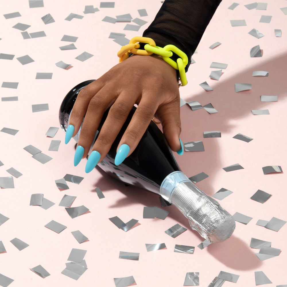 Hand with long sky blue stiletto gel nails resting on a black champagne bottle amid silver confetti on a pale pink background