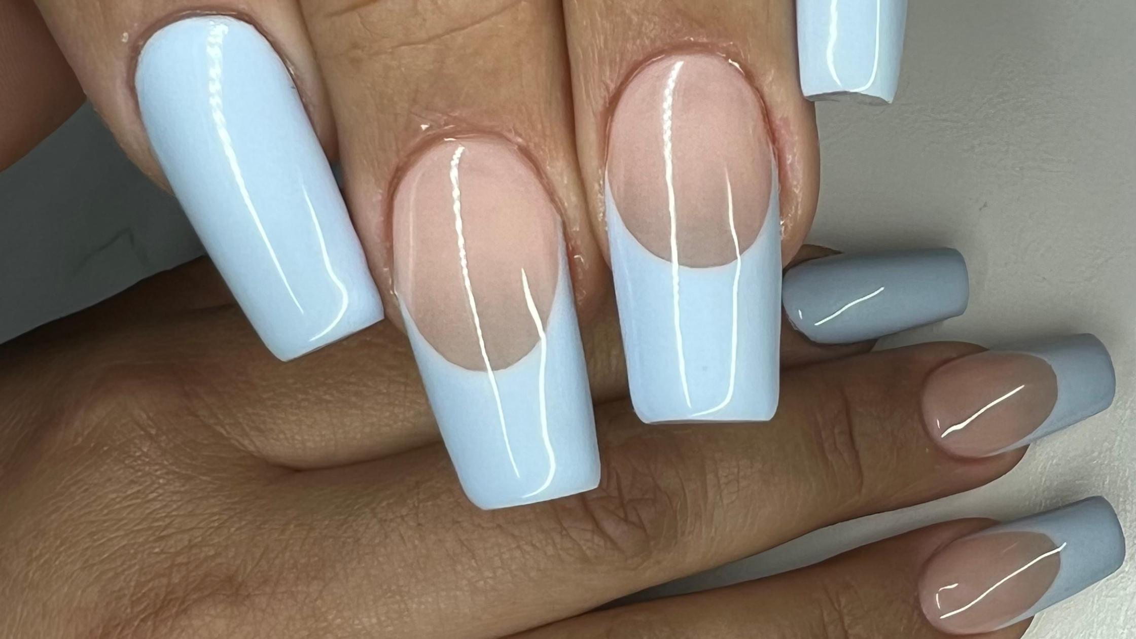 Close-up of long coffin acrylic nails showcasing baby blue opaque polish with nude half-moon negative-space detail on a neutral background